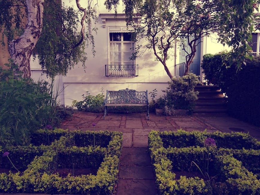 A small, well-maintained garden in Childs Hill with a paved brick pathway leading to a decorative iron bench positioned against a white exterior wall of a house. The garden features neatly trimmed box